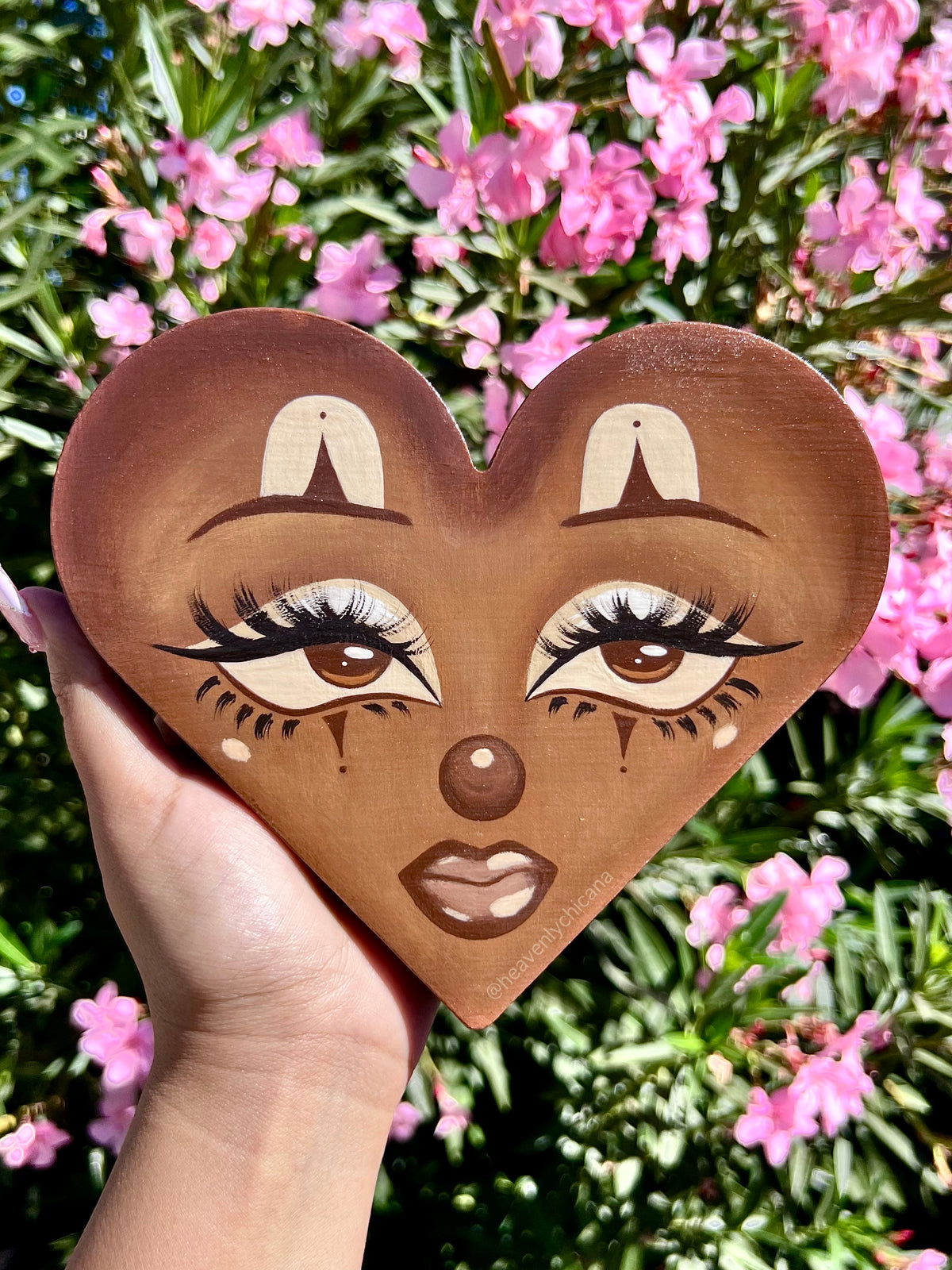 A hand holds a heart-shaped wooden plaque featuring a stylized face with long eyelashes, painted in warm brown tones, set against a background of pink flowers and green foliage.
